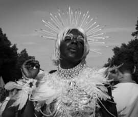 Diva 3 Eine mit Sternkrone, platinblonder Perücke, Perlen-und Federn besetztes Oberteil, stark geschminkte Person posiert lasziv, hebt eine Hand und neigt den Kopf, schaut mit halboffenen Augen in die Kamera. CSD 2023