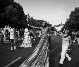 vogue 2 Eine Person mit langem Gewand trägt eine Regenbogenfarbe die Arme weit ausgetreckt. CSD 2023. Eine Frau mit einer Flasche in der Hand streckt die andere Hand zum Berühren aus