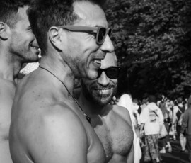 Anthony Drei Männer stehen links, die zwei Vorderen Oberkörperfrei und Sonnenbrillen. Breites Lachen. Eine Person schaut direkt in die Kamera. CSD 2023
