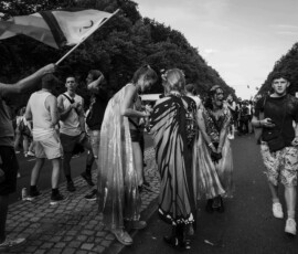 waving, walking, chillin Kamera richtet sich auf Fluchtpunkt. Im Hintergrund weit entfernt das Rote Rathaus schemenhaft. Rechts, eine auf die Kamera zulaufende und blickende Person. Links, eine Person eine Flagge schwenkend, Mitte, lachende und unterhaltende Personen