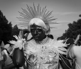 Diva 2 Eine mit Sternkrone, platinblonder Perücke, Perlen-und Federn besetztes Oberteil, stark geschminkte Person posiert lasziv, hebt eine Hand neigt den Kopf runter, schaut mit offenen direktem Blick in die Kamera. CSD 2023