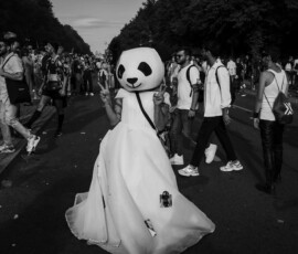 panda Eine mit Panda Kopf und weißer langer Robe steht in Blickrichtung der Kamera und hebt beide Arme hoch, die Finger zu Victory Zeichen geformt. Im Hintergrund Menschenmenge und in der Ferne Fernsehturm sichtbar