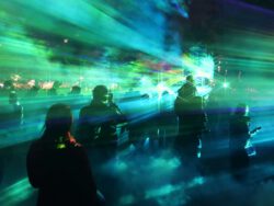 Silhouetten von Menschen vor blau-grünen Lichtstrahlen einer Lasershow bei Nacht, im Hintergrund die beleuchtete Fassade des Humboldtforums in Berlin