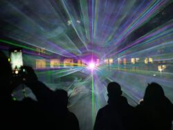 Silhouetten von Menschen vor blau-grünen Lichtstrahlen einer Lasershow bei Nacht, im Hintergrund die beleuchtete Fassade des Humboldtforums in Berlin