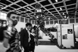 Menschen gehen von links nach rechts. Im Hintergrund ist in Laufschritt von rechts nach links ein Roboter in doppelter Größe